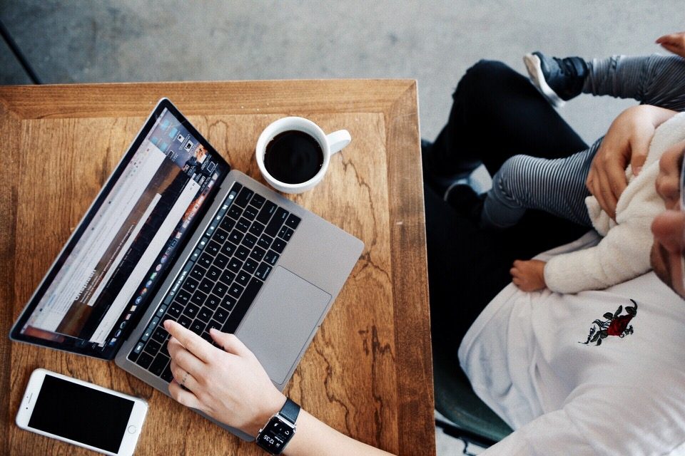 A laptop and a coffee on a wooden desk, with a baby held in someone's arms beside it